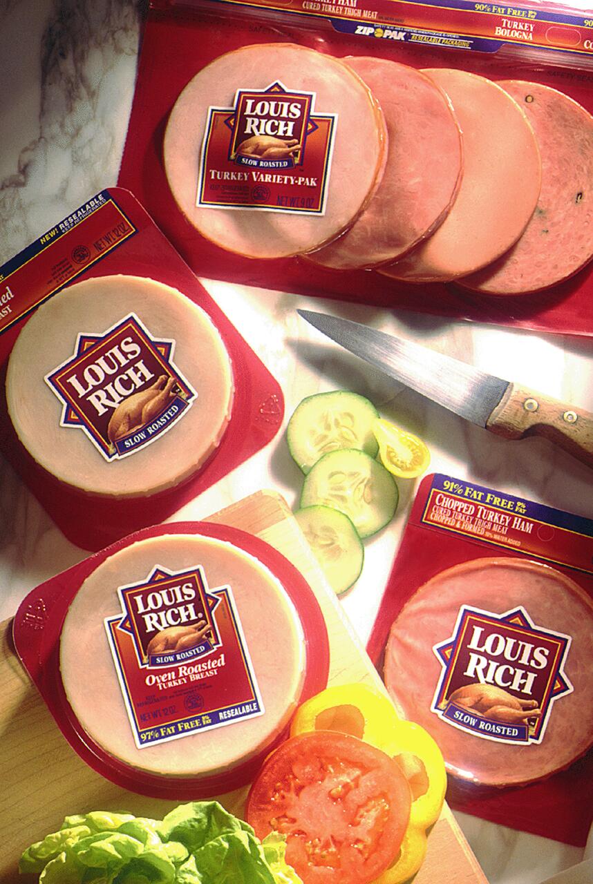 Packaged deli meats labeled "Louis Rich" on a marble surface, surrounded by sliced cucumbers, tomatoes, yellow bell peppers, and lettuce. A knife lies nearby.