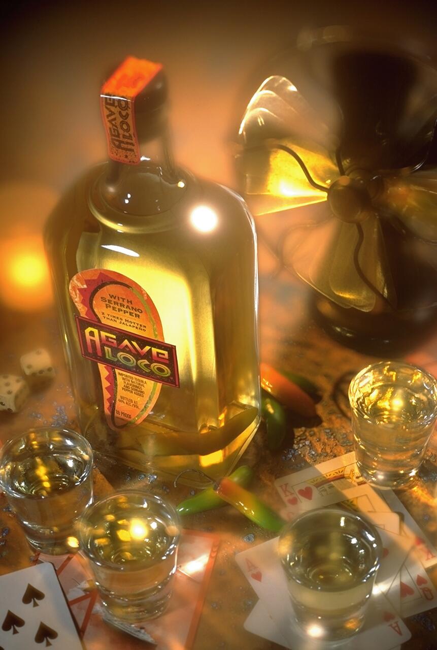 A glass bottle labeled "Agave Loco" with serrano peppers, surrounded by shot glasses filled with liquid, chili peppers, playing cards, dice, and a golden ribbon in warm lighting.