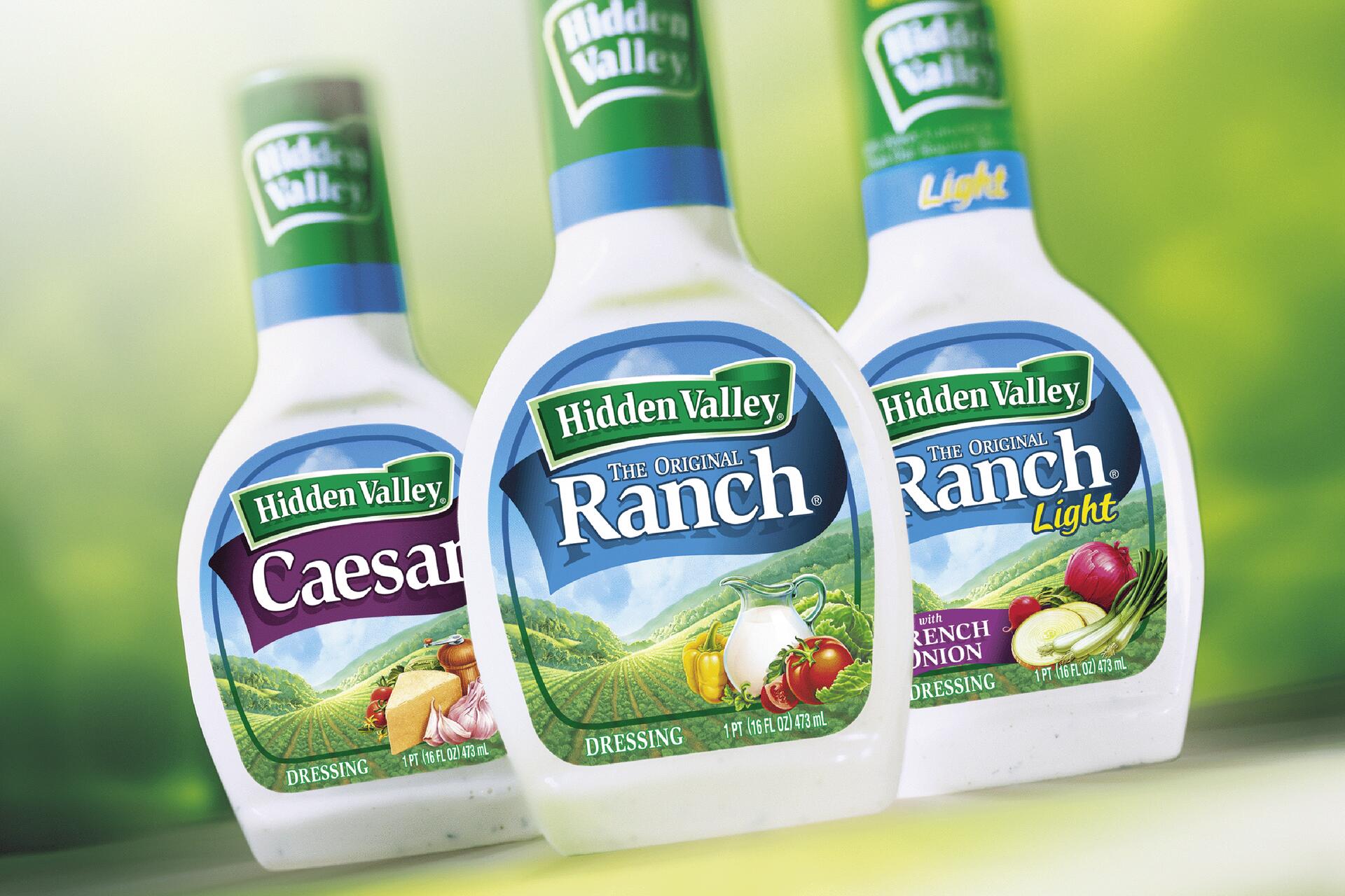 Three bottles of Hidden Valley salad dressing: Caesar, Original Ranch, and Ranch Light. The labels display ingredients and landscape imagery, against a blurred green background.