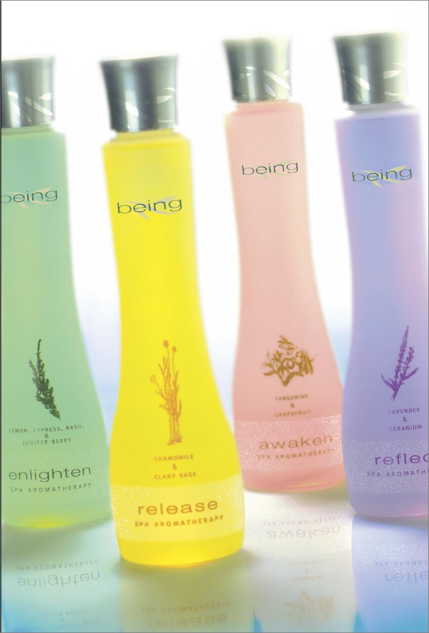 Four colorful spa aromatherapy bottles with silver caps, labeled "enlighten," "release," "awaken," and "reflect." Each bottle is in a different pastel color: green, yellow, pink, and purple, and features botanical illustrations.
