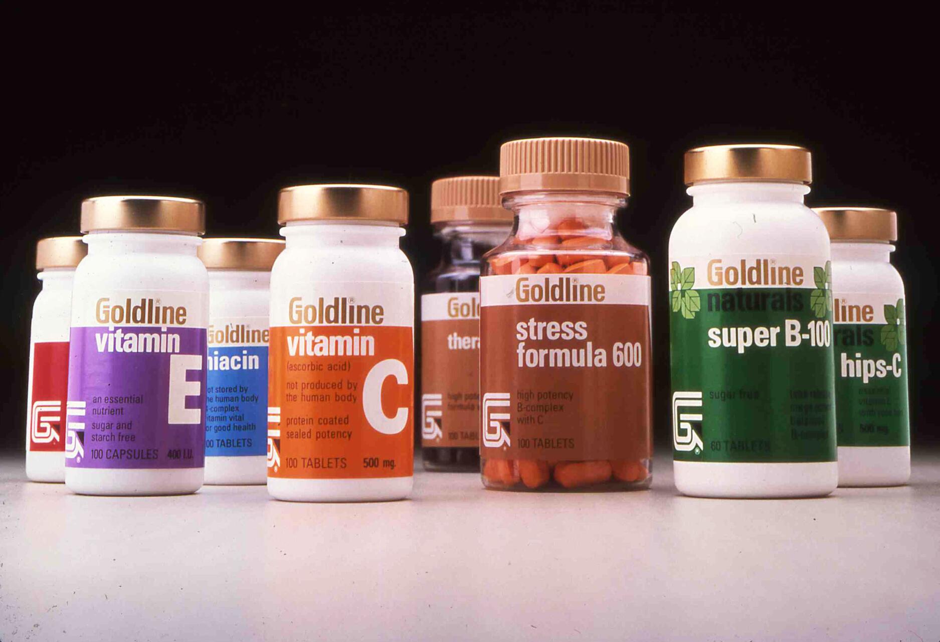 Assorted Goldline vitamin bottles with colorful labels and gold caps, including Vitamin E, C, stress formula, and super B-100, arranged on a light surface against a dark background.