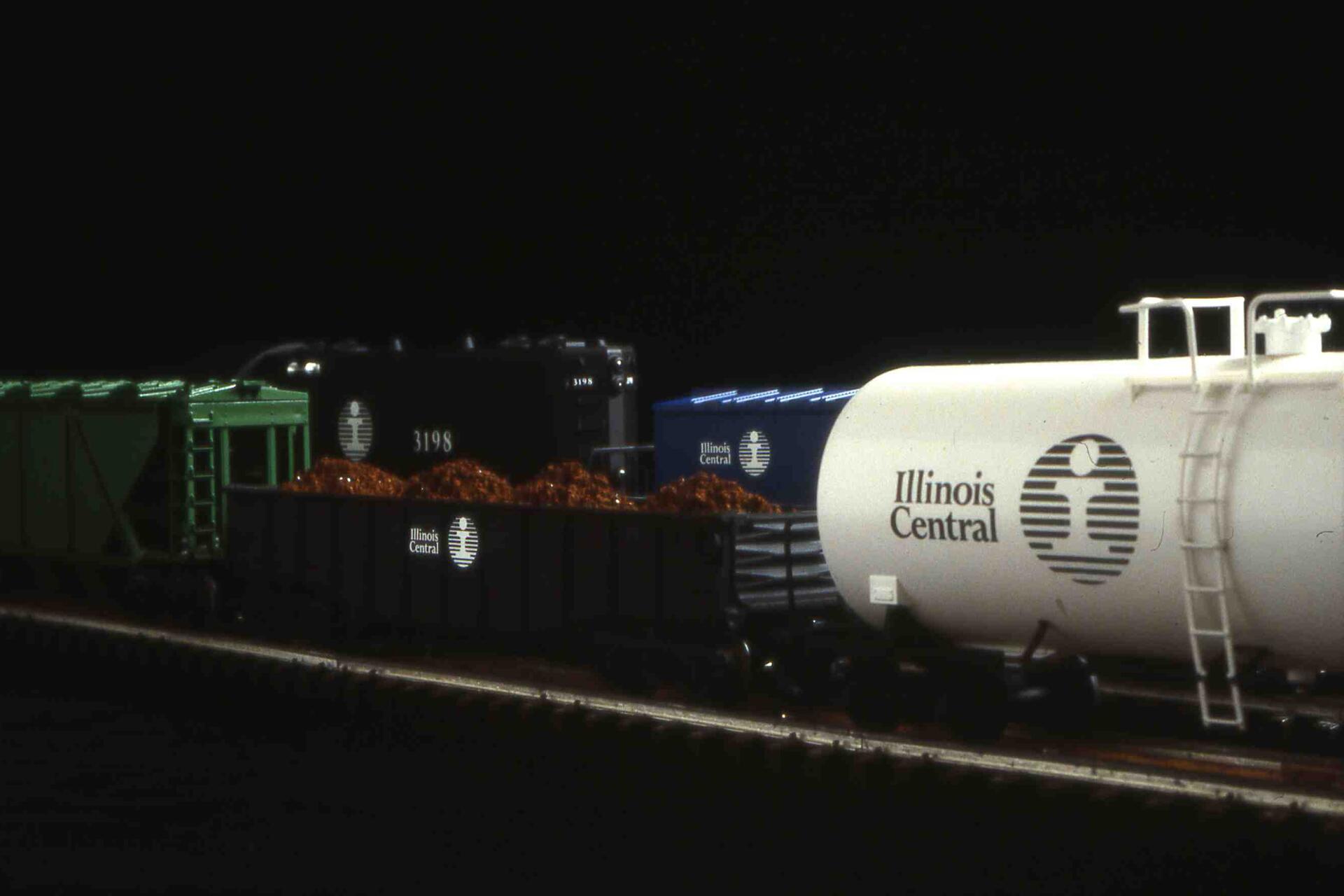 Model train with various colored cars, including a white tanker labeled "Illinois Central," a black cargo car loaded with material, and green and blue cars, set against a dark background.