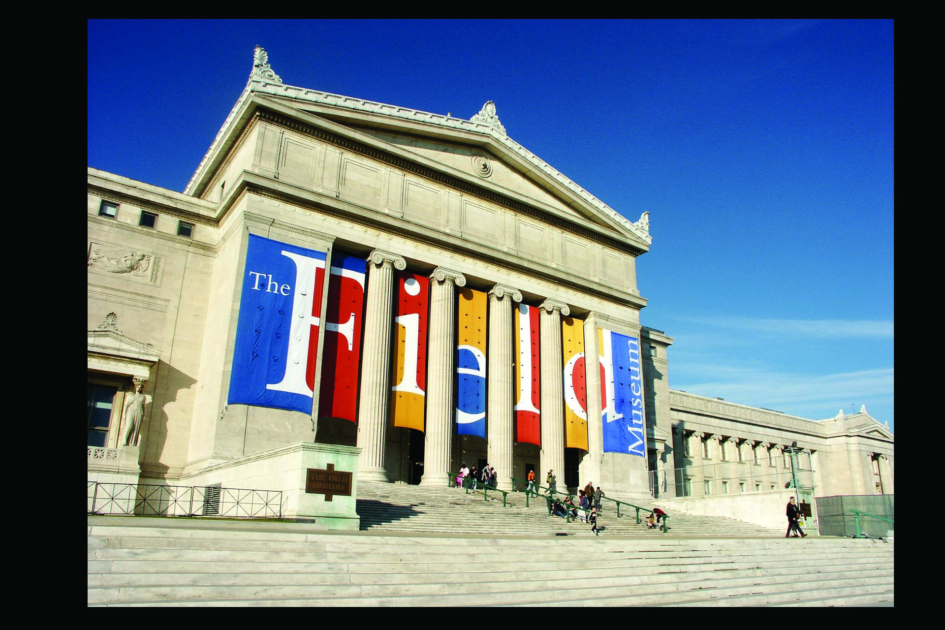 Neoclassical museum building with large, colorful banners hung between columns, people on steps, and a clear blue sky.