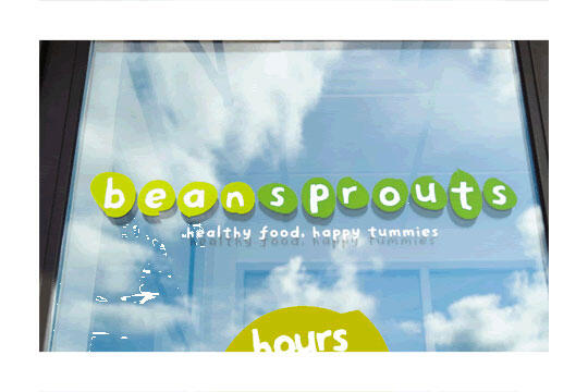 Storefront window with the word "beansprouts" in green, set against a reflective glass showing clouds and blue sky. Slogan below reads "healthy food, happy tummies" in white.