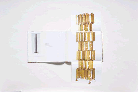 Open book with a pop-up gold architectural structure extending vertically, resembling a modernist building. The left page shows a black and white image and text. White background.
