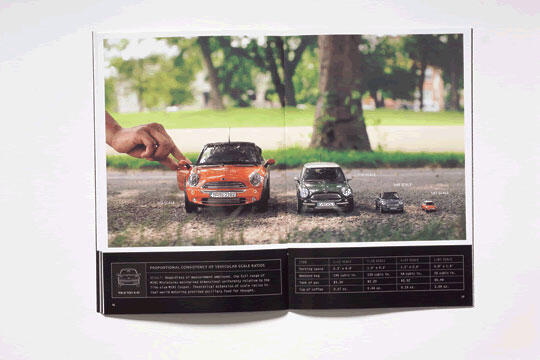 A magazine spread showing a person’s hand touching a series of progressively smaller toy cars lined up on a gravel path, with a park setting in the background.