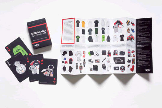 Box of Mini Brand playing cards next to a few scattered cards featuring watches and keychains. An unfolded brochure displays various merchandise, including clothing and accessories, on a white surface.