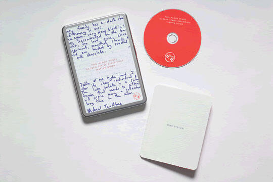 A tin box with handwritten text on the lid, a red CD, and a small white booklet laid on a light surface.