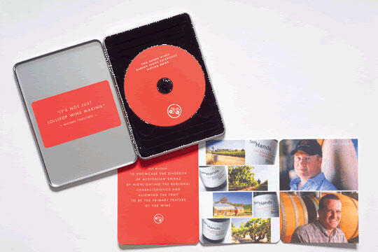 Open tin box with a red disc inside, alongside a red card with writing. Next to them, a folded leaflet showing vineyard images and close-ups of two people.
