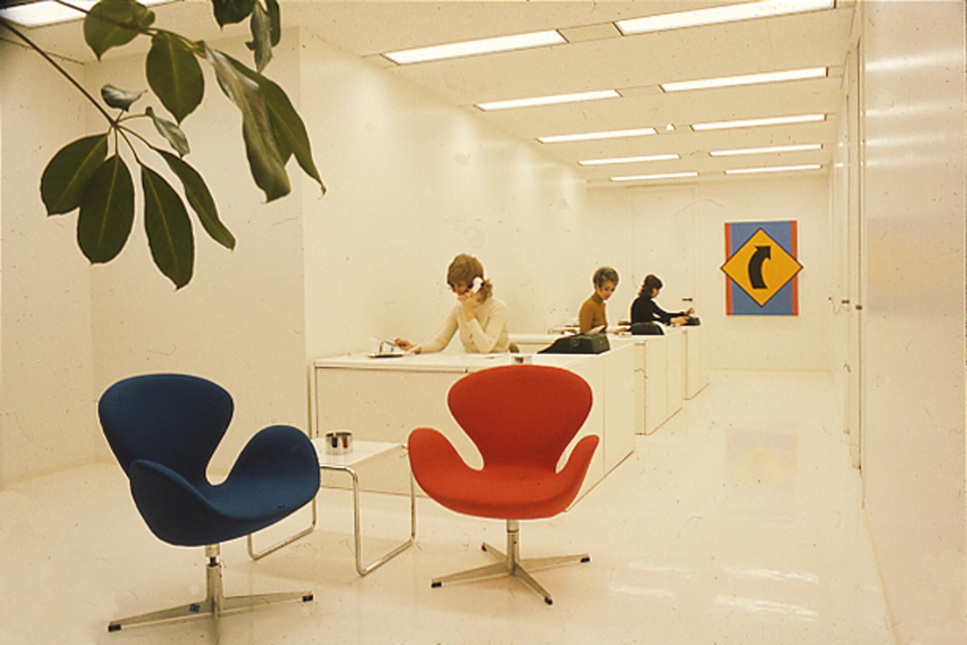 Minimalist office with white walls, a blue and a red mid-century modern chair, and three people working at desks in the background. A colorful abstract artwork hangs on the wall. A leafy plant is in the foreground.