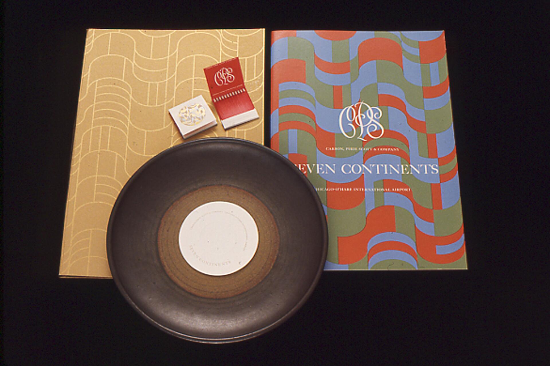 A decorative arrangement featuring a brown plate with a central white circle, a gold-patterned book or folder, and a colorful, wavy-patterned brochure labeled "Seven Continents." Two matchbooks with ornate designs are on top.