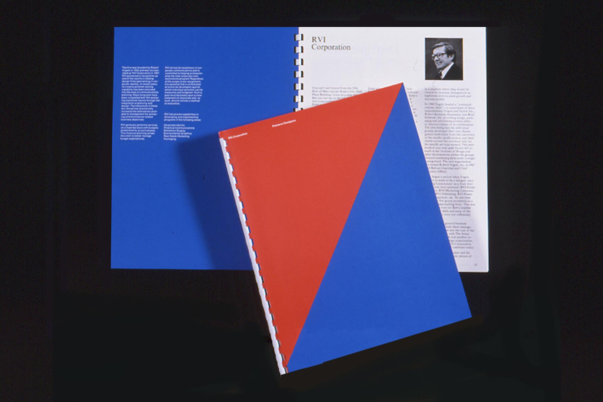 Two spiral-bound booklets, one with a red cover and blue triangle, the other open showing text and a portrait. Both rest on a blue surface.