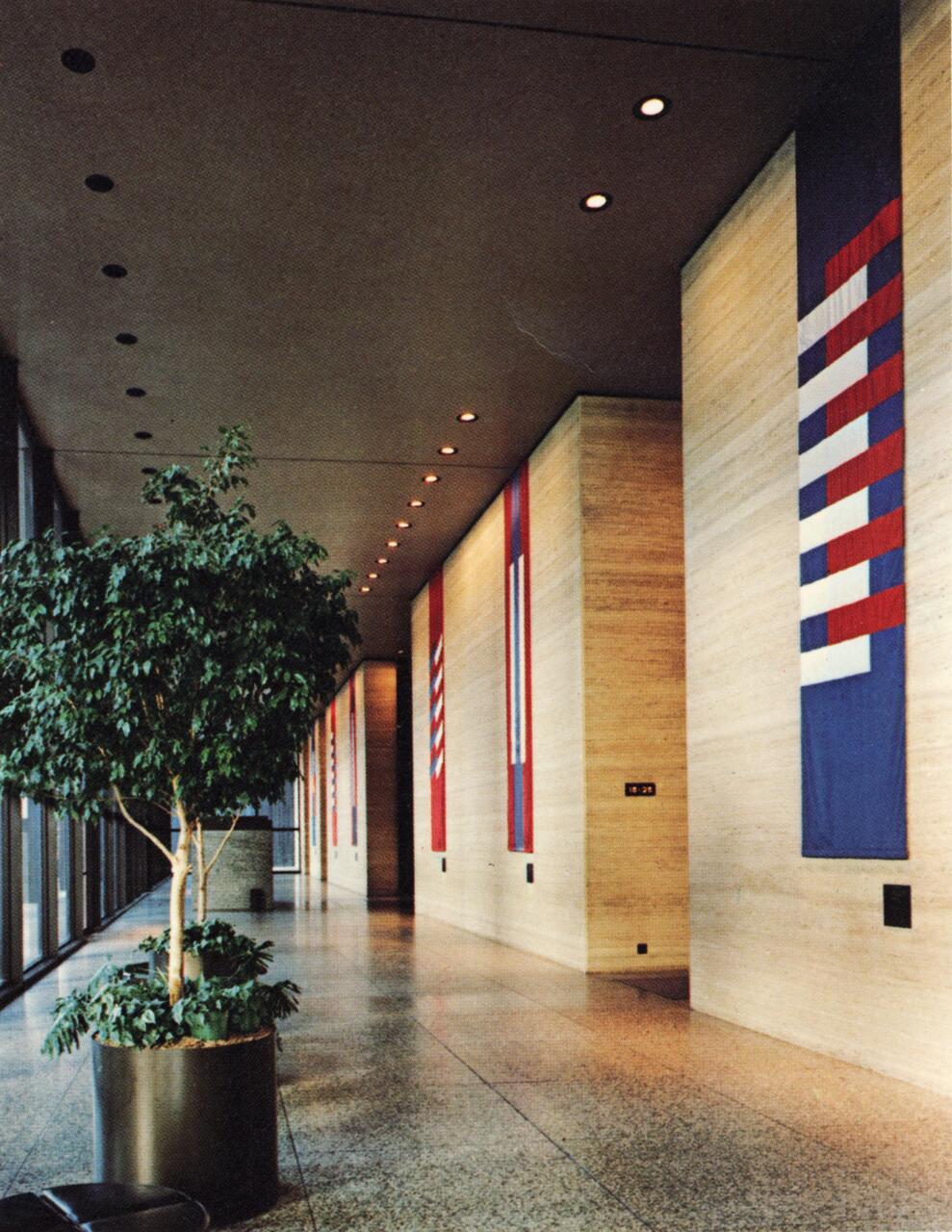 Hallway with large beige walls, decorated with vertical banners of geometric red, white, and blue patterns. A potted tree sits on the left, natural light streams through the glass wall, and recessed ceiling lights illuminate the space.