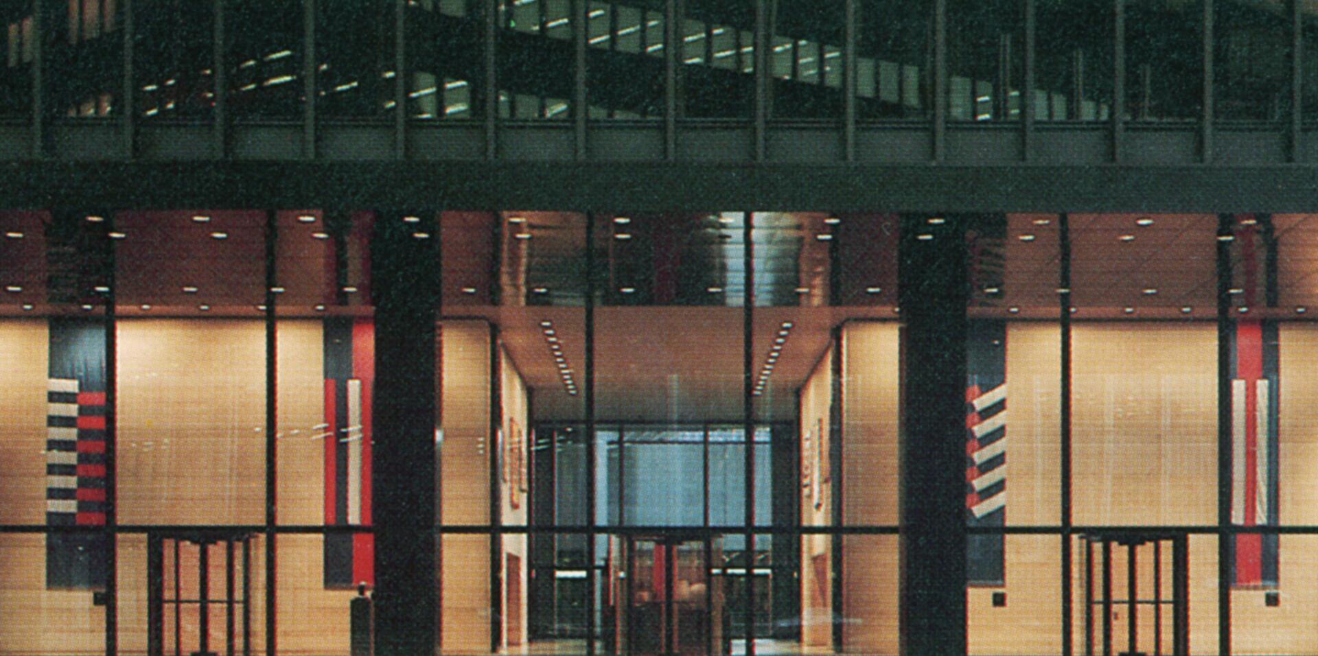 Modern building lobby with glass facade, interior brightly lit. Tall vertical banners in red, blue, and white hues decorate the walls. Large glass doors lead inside, surrounded by reflective walls and a ceiling with recessed lights.