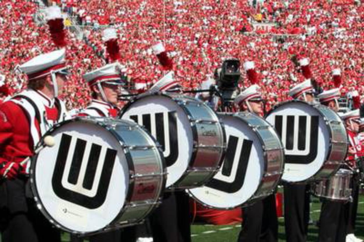 Marching band members in red and white uniforms play large bass drums with black 'W' logos, set against a backdrop of a crowd dressed in red.