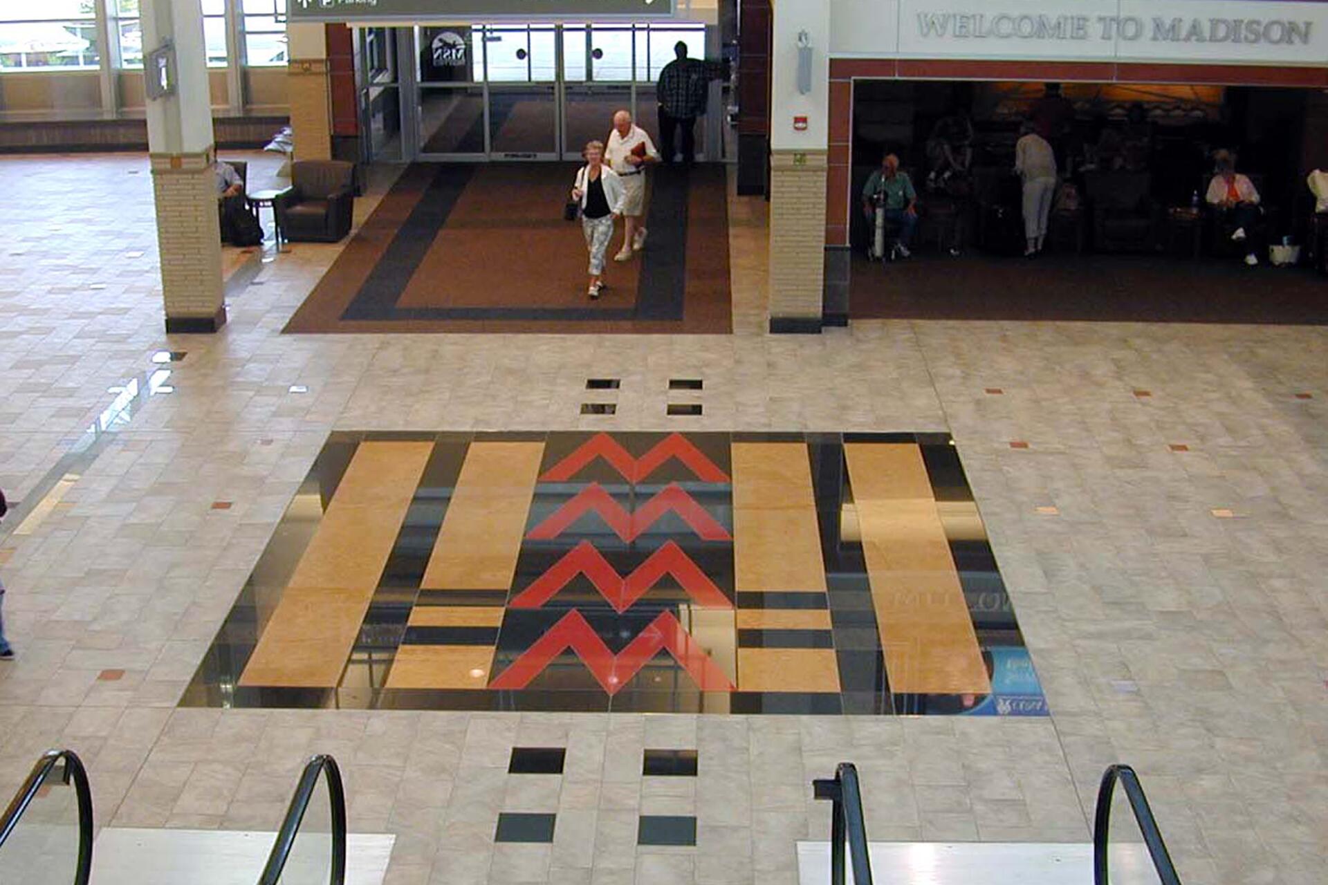 Spacious airport terminal with tiled floor design in red, black, and beige. Escalators lead to the area, where people walk and sit. Large windows provide natural light.