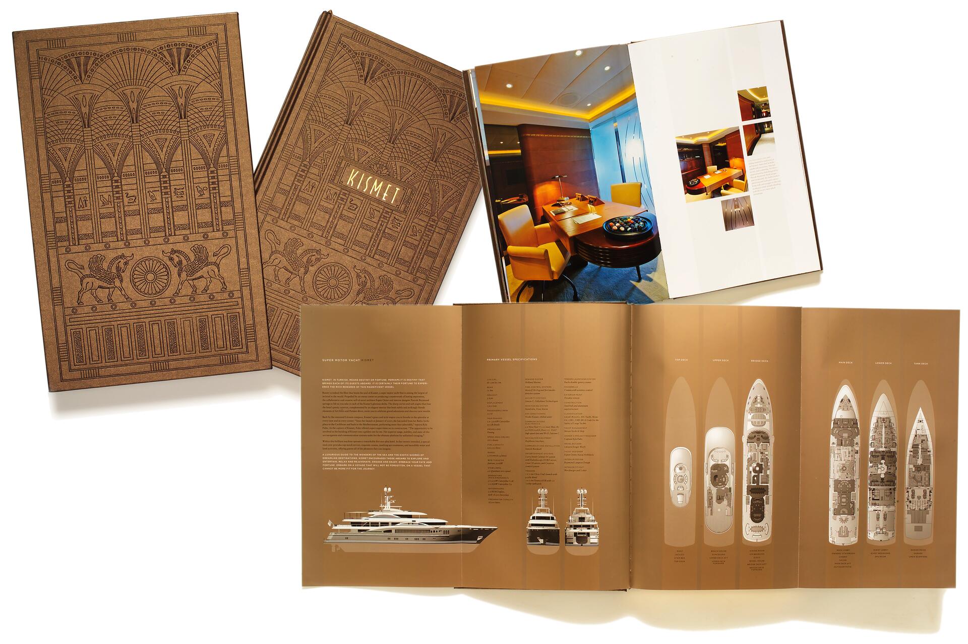 Two brown hardcover books with intricate designs, labeled "Kismet." One book is open, showing a luxurious yacht interior photo on glossy pages. Below, unfolded pages detail yacht specifications, including illustrations of yacht cross-sections and a side profile drawing.