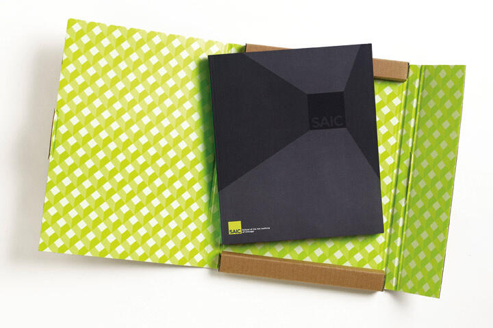 Black book with "SAIC" on the cover, placed in an open cardboard box lined with green and white checkered pattern.