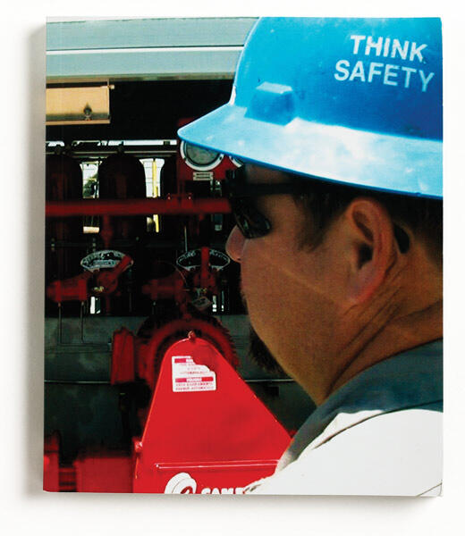 Man wearing a blue hard hat labeled "Think Safety" and sunglasses, standing near red industrial equipment.