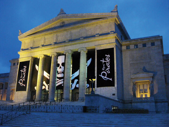 Classical building at dusk with large pirate banners featuring skulls and swords hanging between columns.