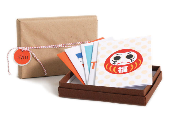 A small brown box containing colorful folded note cards with one featuring a cartoonish Daruma doll. A gift wrapped in brown paper and twine is in the background.