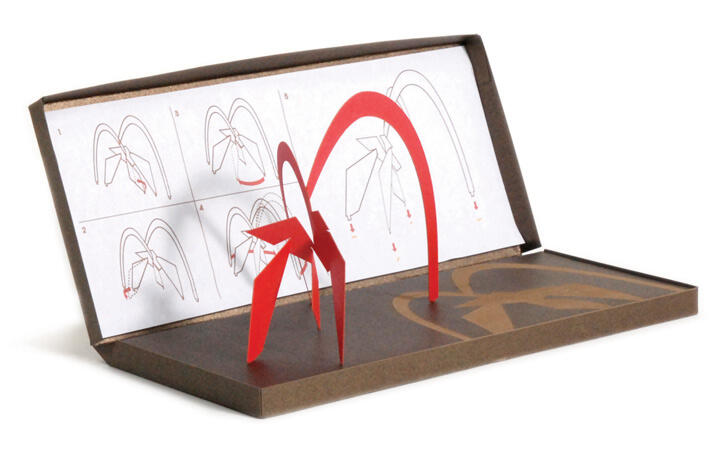 Red paper sculpture resembling an arch and abstract figure inside an open brown box. Background features folding instructions.