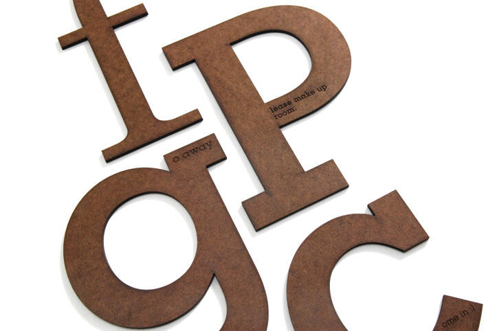 Brown wooden alphabet letters on a white background, with small engraved text on some letters like "o away" and "please make up room."