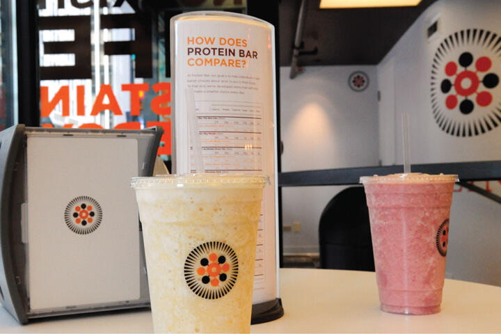 Two smoothies, one yellow and one pink, on a table with a nutritional comparison chart and branded signage in the background.