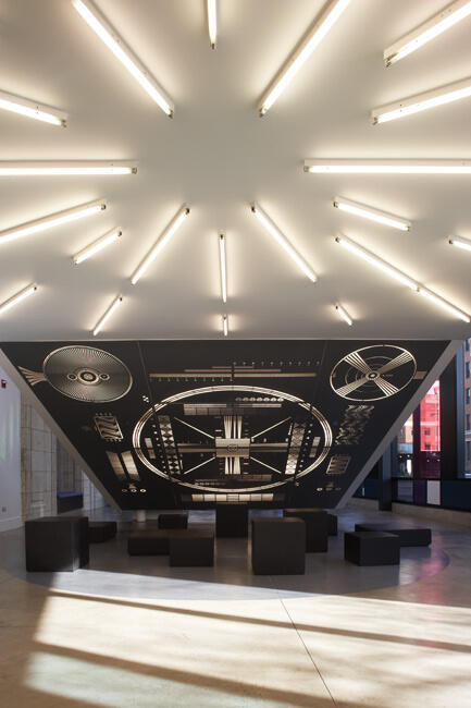 Modern interior with an inverted pyramid ceiling, featuring black and white geometric patterns and test chart designs. Rows of bright white fluorescent lights radiate outward. Minimalist black seating is arranged on the light-colored floor.