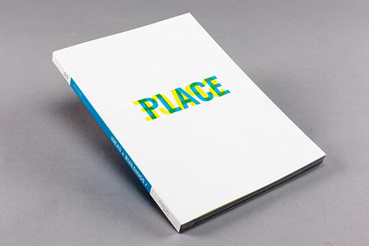 A book with a white cover displaying the word “PLACE” in colorful, overlapping letters. The spine has a blue strip with text. The book is photographed against a gray background.