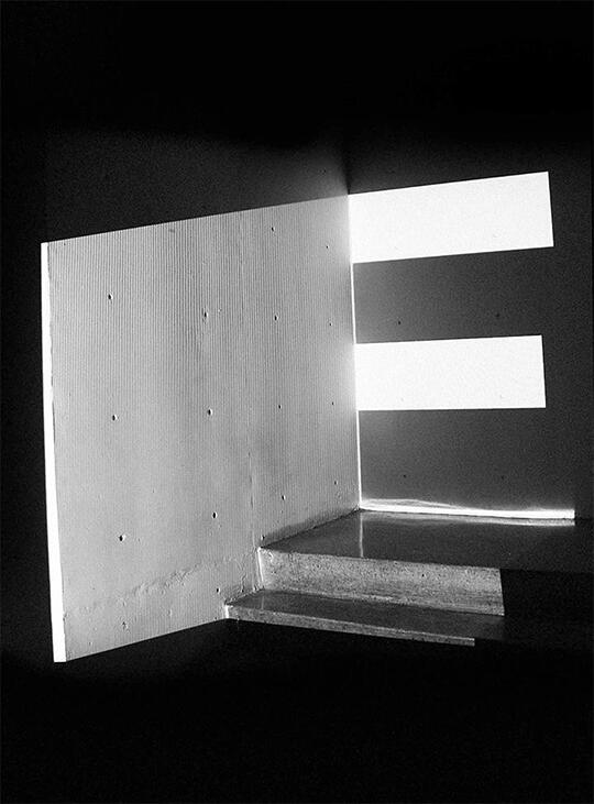 Black and white photograph of a wall and stairs, with strips of light casting geometric shadows, creating a minimalist and abstract pattern.