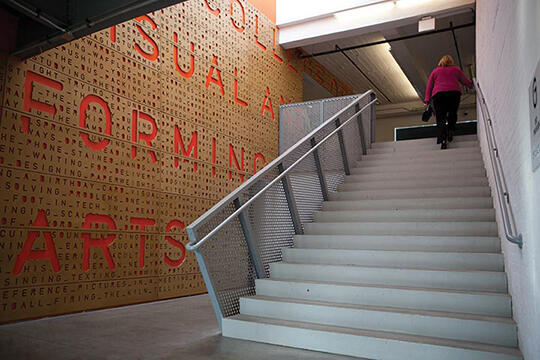 A person in a pink sweater climbs a set of stairs in a building. The wall to the left features large red letters and words in varying fonts. The stairway leads to a brightly lit area above.