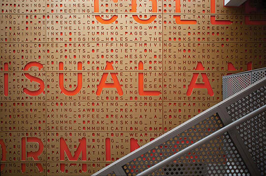 A perforated metal staircase in front of a wall with large red letters spelling "VISUAL ART" and smaller text in a decorative pattern.