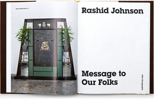 Open book displaying an artwork featuring a large, dark panel with a central rectangular section and a small face above it. Two green plants flank the panel. Opposite page reads "Rashid Johnson, Message to Our Folks."