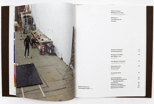 Open book showing an artist's studio on the left page, with a person standing next to a large dark canvas on the floor. Shelves and art supplies fill the background. The right page lists text, possibly a table of contents, with itemized topics and page numbers.