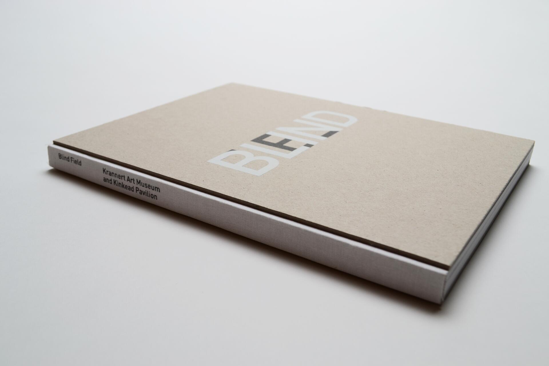 Tan hardcover book titled “BLEND” with white spine text “Blind Field, Krannert Art Museum and Kinkead Pavilion,” on a white surface.