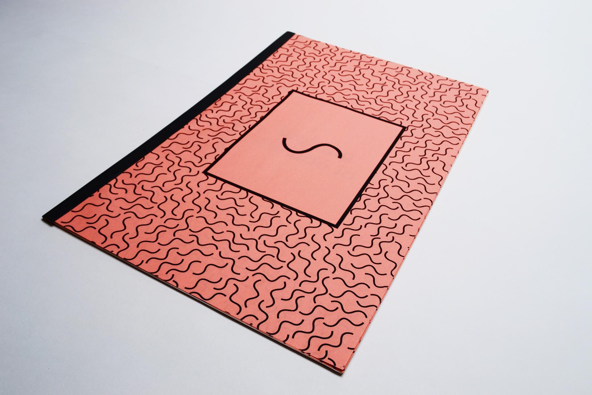 Orange notebook with wavy black lines, a black spine, and a large central rectangle featuring a single 'S' in the middle.