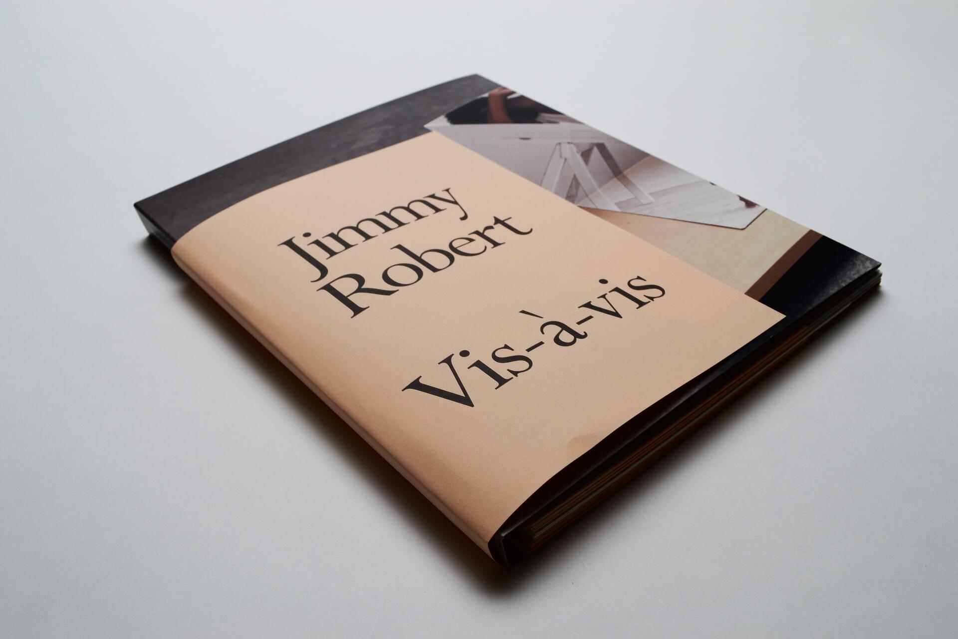A book titled "Vis-à-vis" by Jimmy Robert, with a beige cover and a partial image of papers on a surface. The book is angled on a white background.