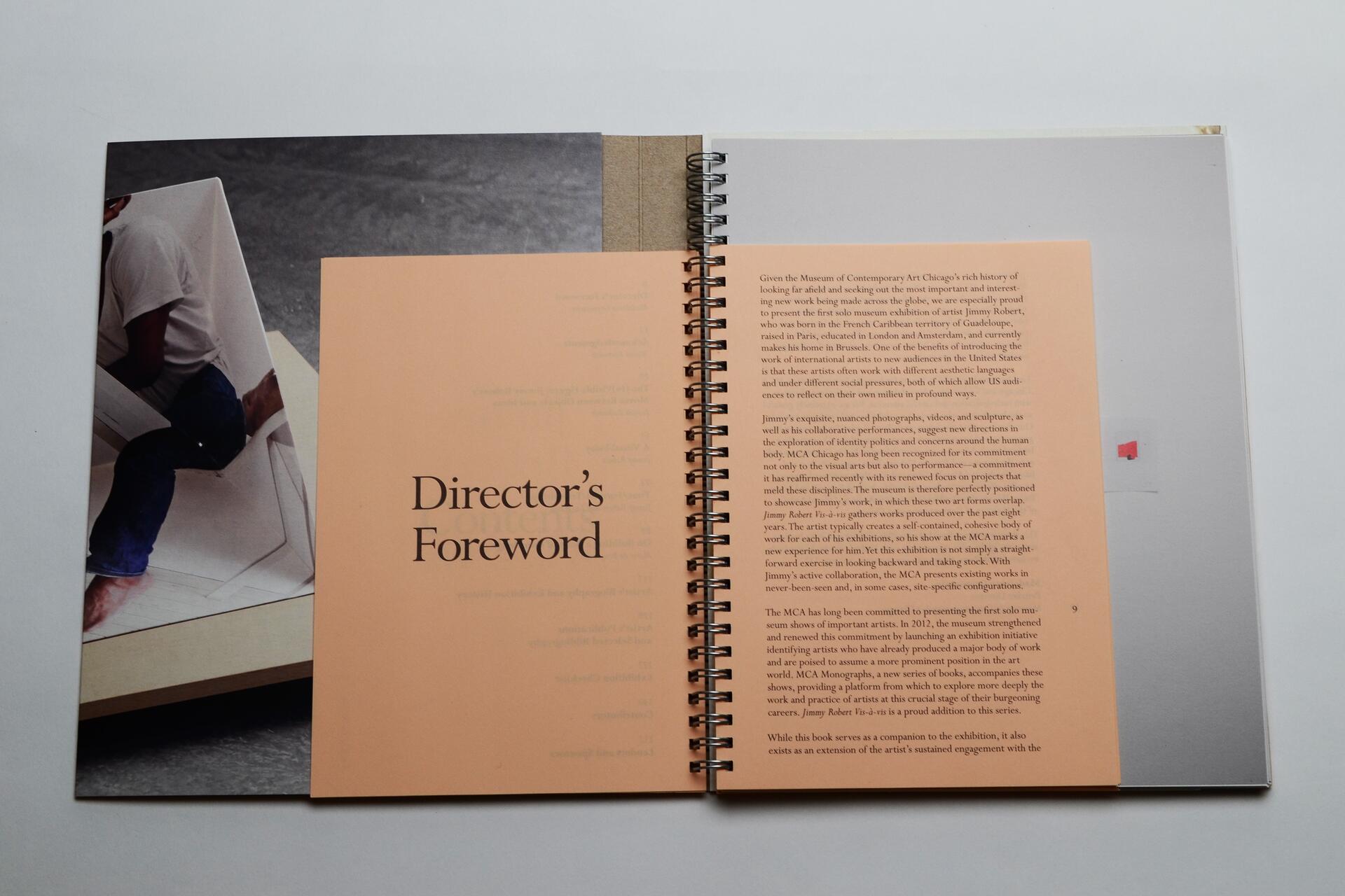 Open book with a peach-colored page titled "Director’s Foreword" on the left and a page of text on the right. The left side partially shows a photograph of a person sitting on a geometric chair, wearing jeans and a white shirt.