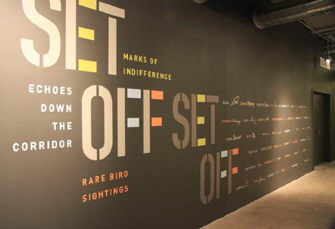 Text-filled corridor wall with large phrases like "SET OFF," "ECCOES DOWN THE CORRIDOR," and "RARE BIRD SIGHTINGS." The words are in varied colors with smaller names or texts aligned along the wall in a decorative pattern.