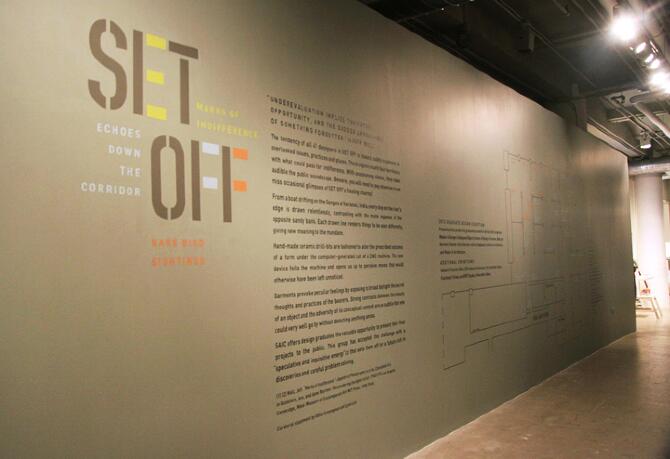 Exhibit wall with text and a blueprint drawing, in an industrial gallery setting. The text includes titles and descriptions of art installations. The lighting is warm, and the floor is polished concrete.