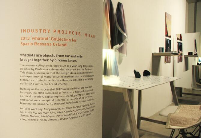 Exhibition space featuring a wall with text detailing the 2013 ‘whatnot’ collection in Milan. A wooden desk with geometric objects and a modern chair is nearby. Art pieces and hanging drapes are visible on the adjacent wall, lit by soft lighting.
