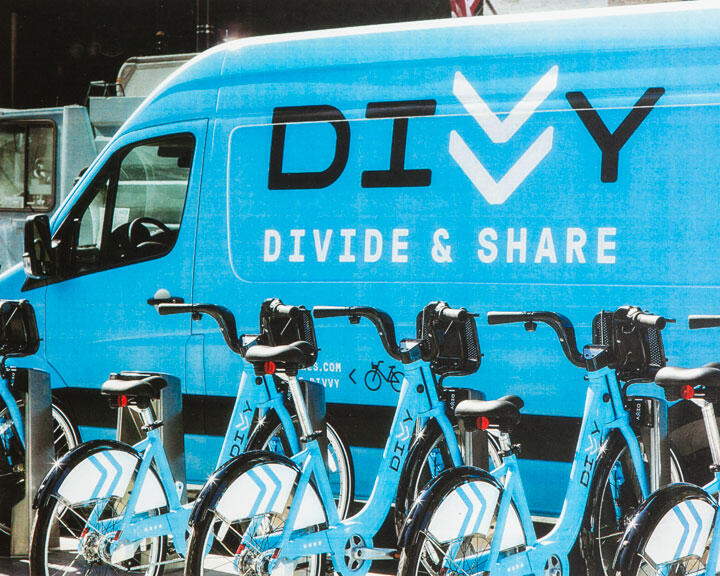 Blue van with "Divvy" and "Divide & Share" text, parked next to a row of blue rental bicycles.