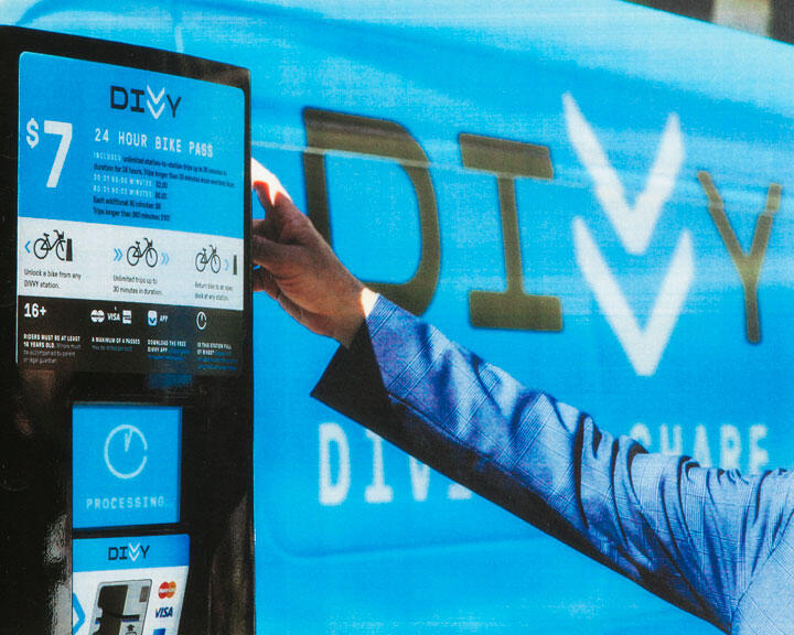 Close-up of a person's arm in a blue suit sleeve reaching toward a Divvy bike rental kiosk. The background features a large blue Divvy logo.