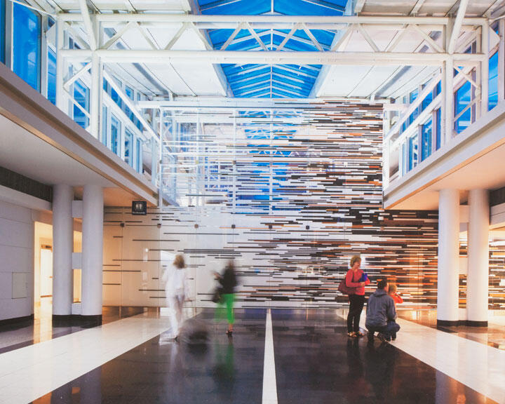 Spacious modern atrium with a high glass ceiling, abstract striped wall design, and people walking or talking. Bright lighting and white pillars create an open, airy atmosphere.