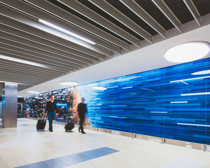 Two individuals pulling luggage walk past a modern airport corridor with a vibrant blue and orange digital art mural. The ceiling features a grid-like design with circular lights, and the floor is tiled in neutral tones.