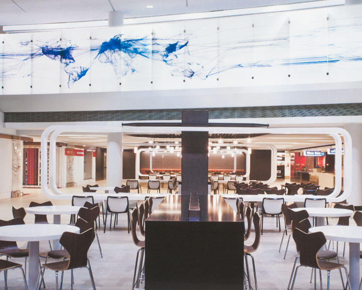 Spacious modern cafeteria with white tables and black chairs, a long counter in the center, and abstract blue artwork on the wall above.
