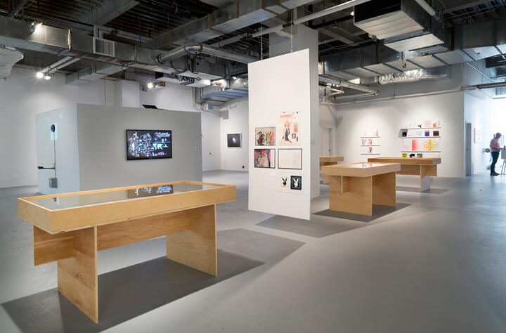 Art gallery interior with modern exhibits. Minimalist tables display art under glass. Walls adorned with framed artworks. Industrial ceiling with exposed ductwork, soft lighting, and a person in the background near vibrant pieces.