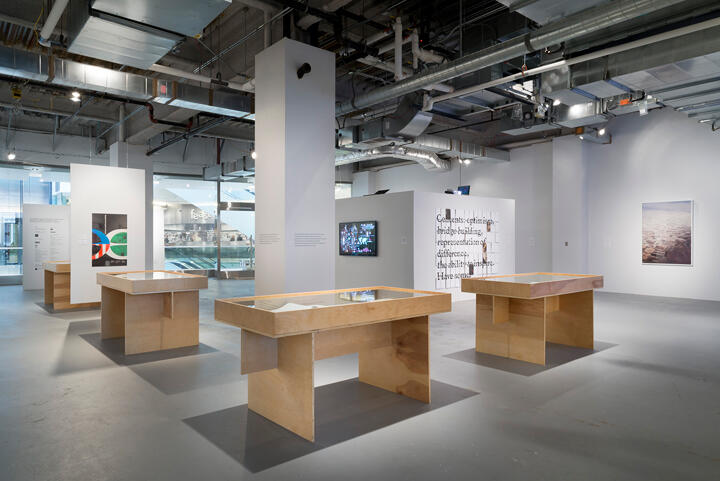 Art gallery interior with four wooden display tables under industrial ceiling. Walls showcase various artworks, including colorful prints and media screens. Large windows allow natural light, creating a spacious, modern atmosphere.