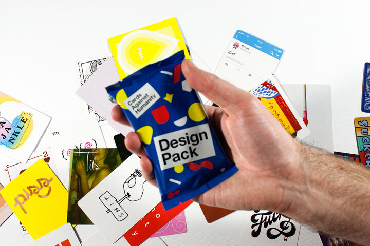 A hand holding a colorful "Design Pack" against a background of various graphic and design cards scattered on a white surface.